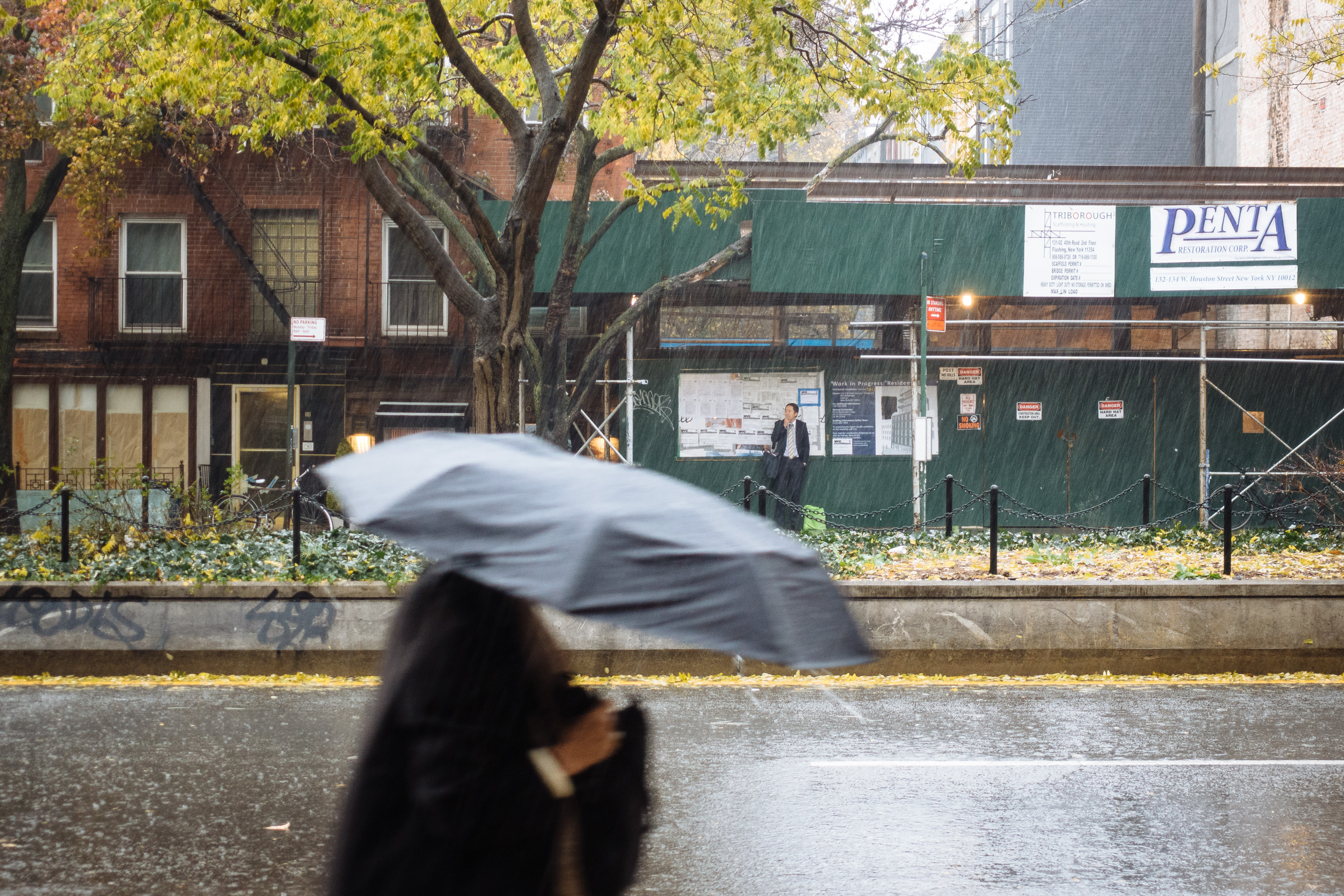A woman under an umbrella walks along Houston Street, a man smokes a cigarette under a scaffold