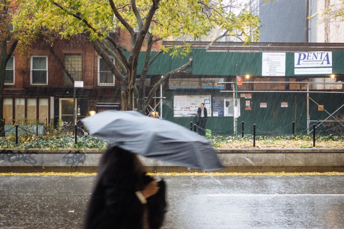 A woman under an umbrella walks along Houston Street, a man smokes a cigarette under a scaffold